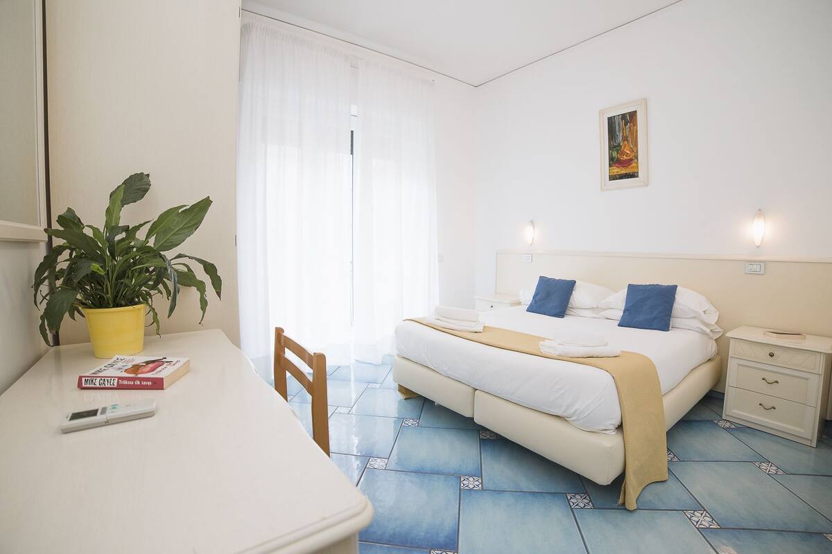 Habitación de hotel con una cama, mesitas de noche, un escritorio con una planta y un libro, y una ventana con cortinas.