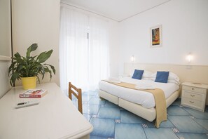 A hotel room with a bed, bedside tables, a desk with a plant and a book, and a window with curtains. - Hotel De Rosa (Maiori)