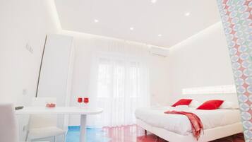 A modern hotel room with a white bed, red pillows, a small table with two glasses, and a white chair.