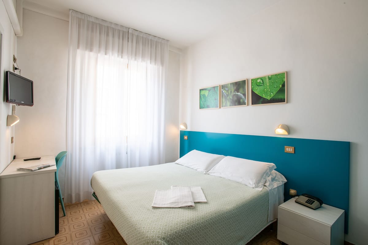 Quarto de hotel com cama, escrivaninha, cadeira, televisão e mesa de cabeceira com telefone.