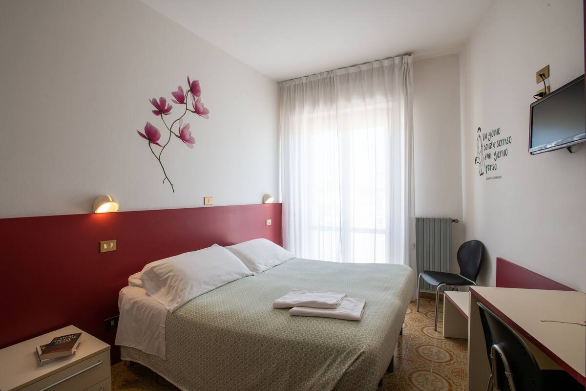 Quarto de hotel com cama, escrivaninha, cadeira, televisão e parede com um desenho floral.