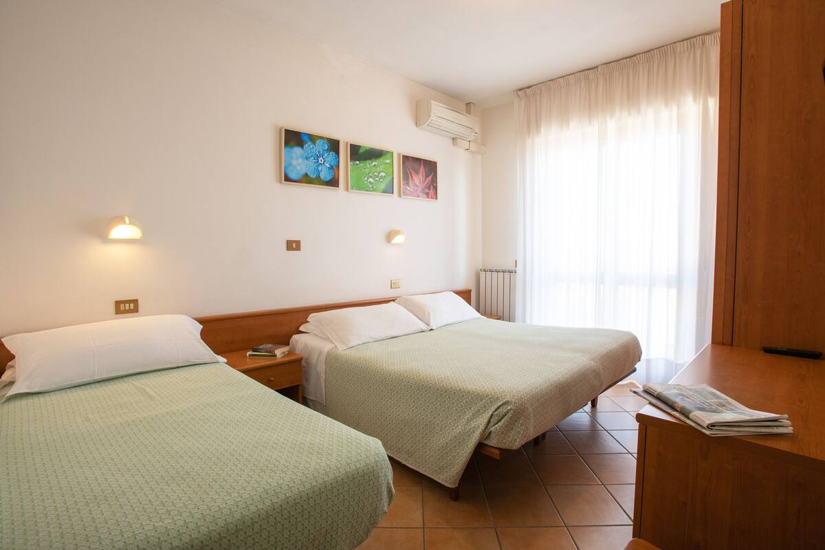 Quarto de hotel com duas camas, uma escrivaninha de madeira, janela com cortinas e obras de arte na parede.