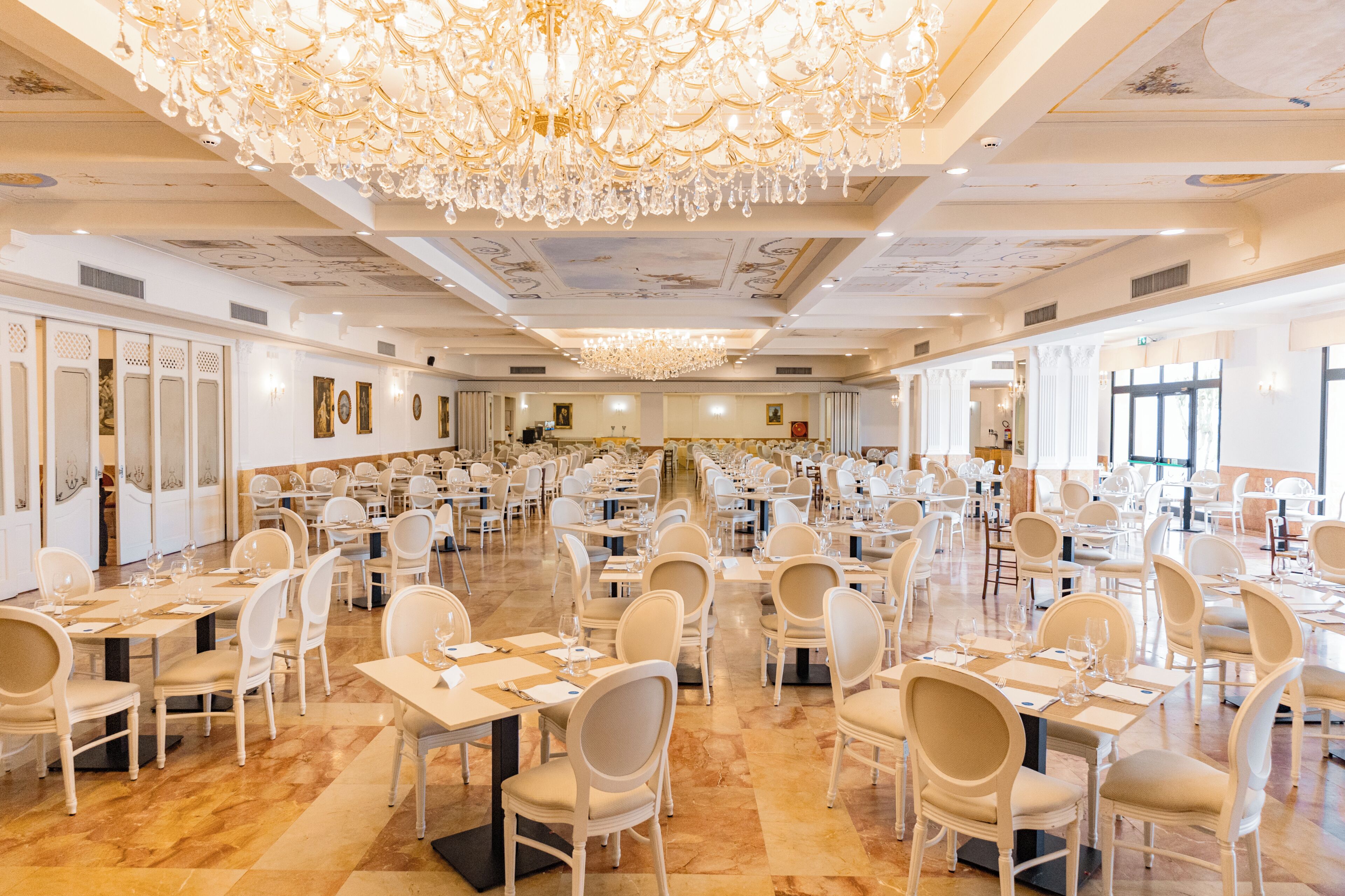 A large banquet hall with numerous tables and chairs, a grand chandelier, and a high ceiling with intricate designs.
