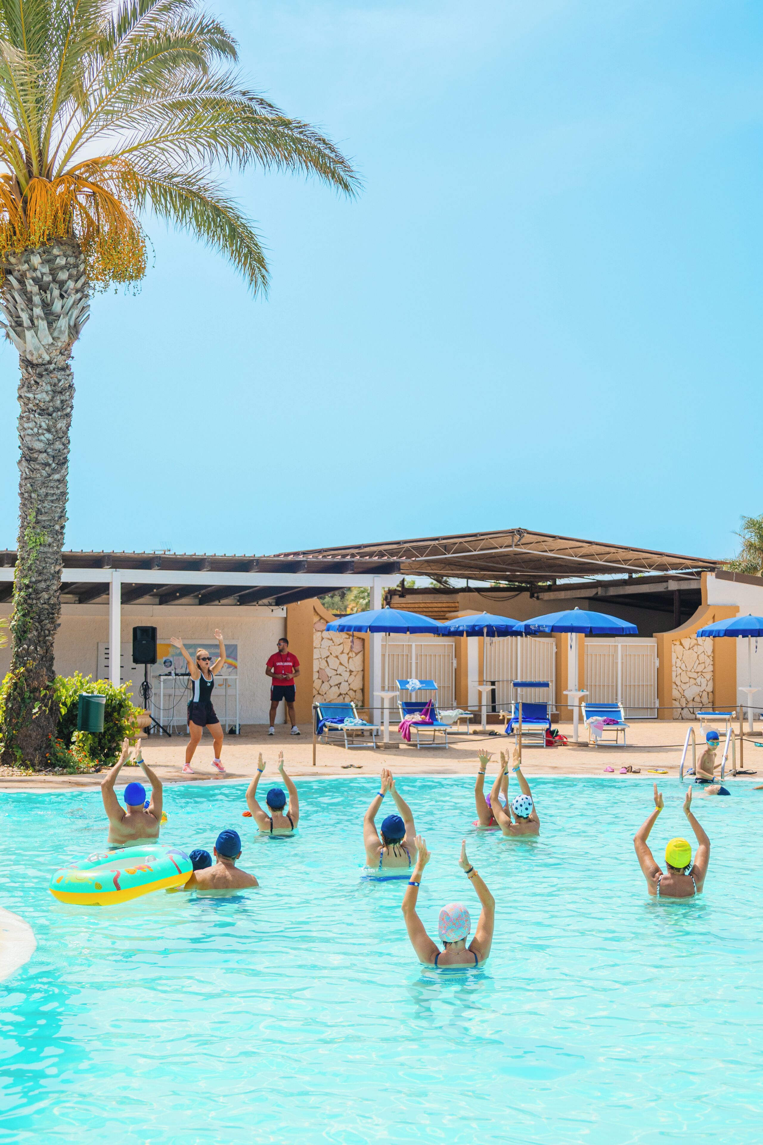 A swimming pool with people playing, palm trees, and umbrellas.