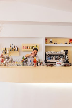 A bar area with a variety of bottles and glasses, a coffee machine, and a counter with a person preparing a drink. - Delfino Beach Hotel (Marsala)