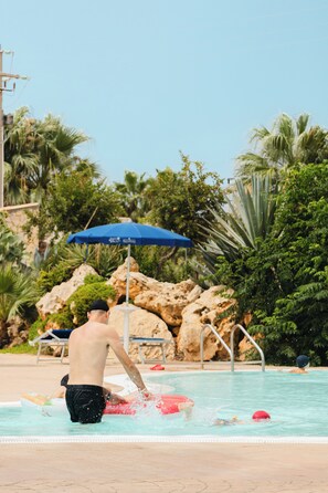 A swimming pool with a person splashing water, surrounded by palm trees and a rocky backdrop. - Delfino Beach Hotel (Marsala)