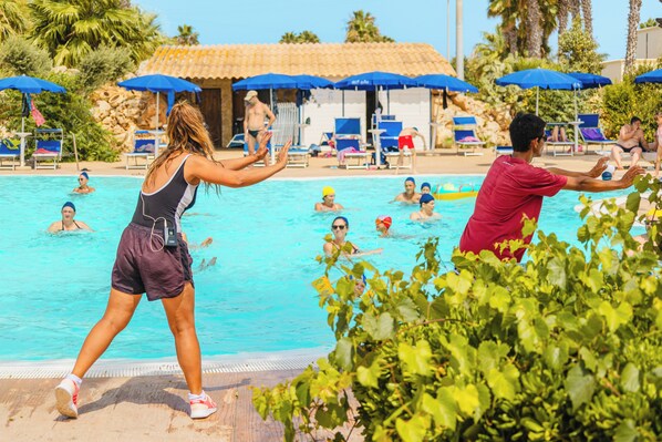 Zona de piscina con gente nadando y tomando el sol, rodeada de palmeras y sombrillas azules.