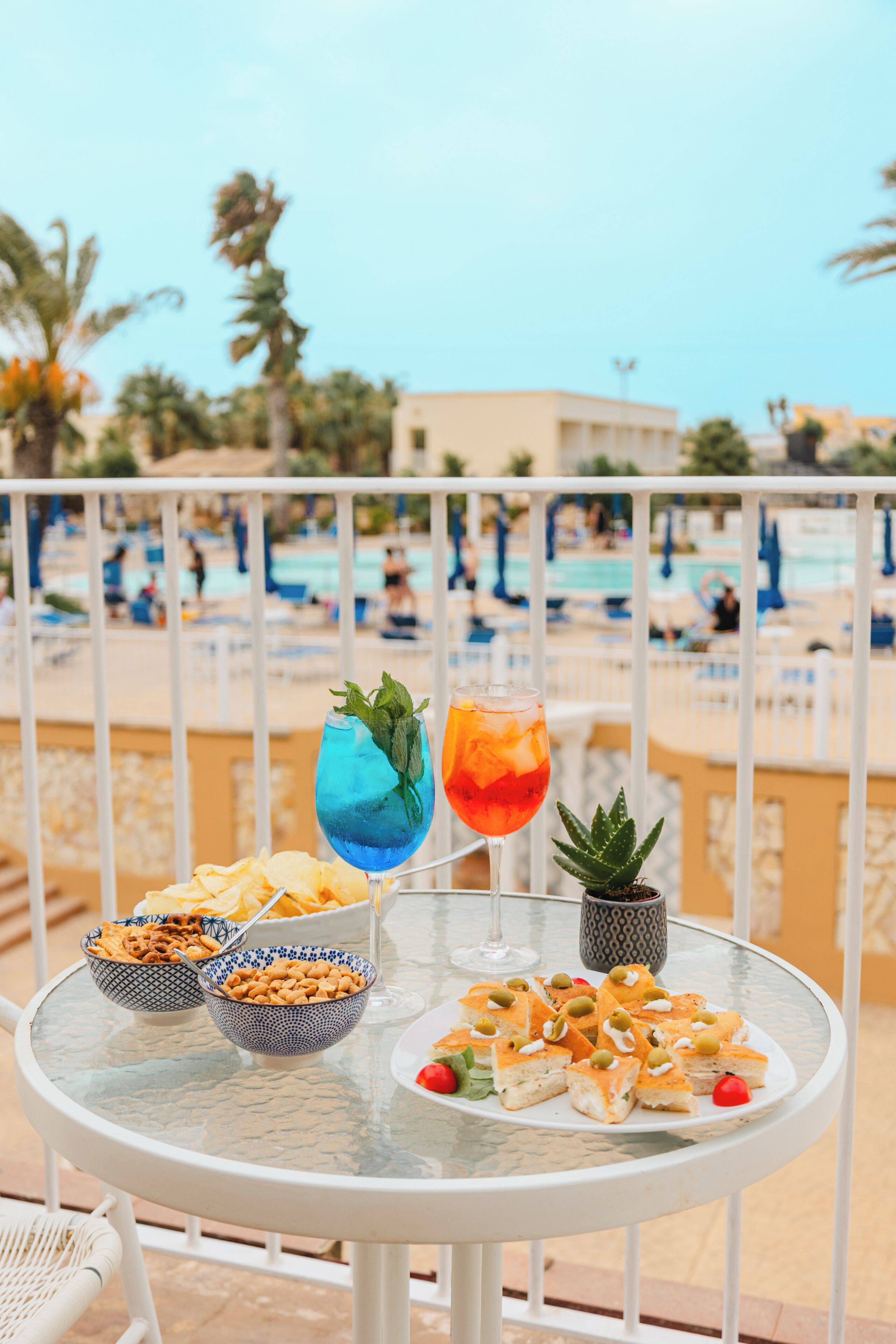Un tavolo con snack, bevande e una piccola pianta su un balcone con vista su una zona piscina.