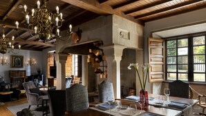 A dining area with a wooden table set for two, surrounded by upholstered chairs. A chandelier hangs from the ceiling, and there are windows with a view of greenery outside.