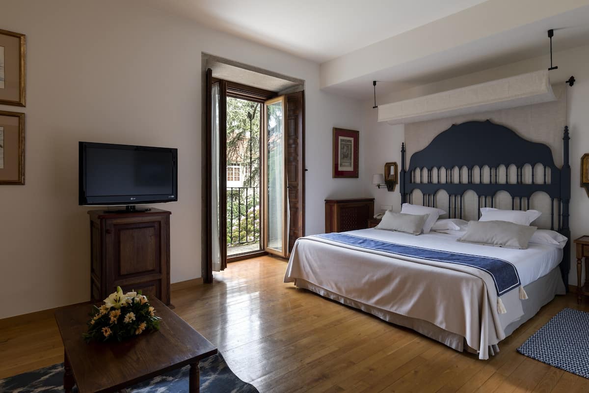 A bedroom with a bed, a television on a wooden cabinet, a small table with flowers, and a door leading to an outdoor area.