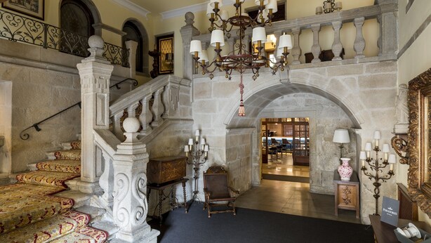 A grand staircase with ornate railings and a chandelier, leading to a lobby area with a stone fireplace and a seating area.