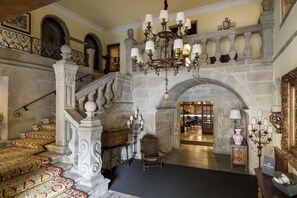 A grand staircase with ornate railings and a chandelier, leading to a lobby area with a stone fireplace and a seating area.