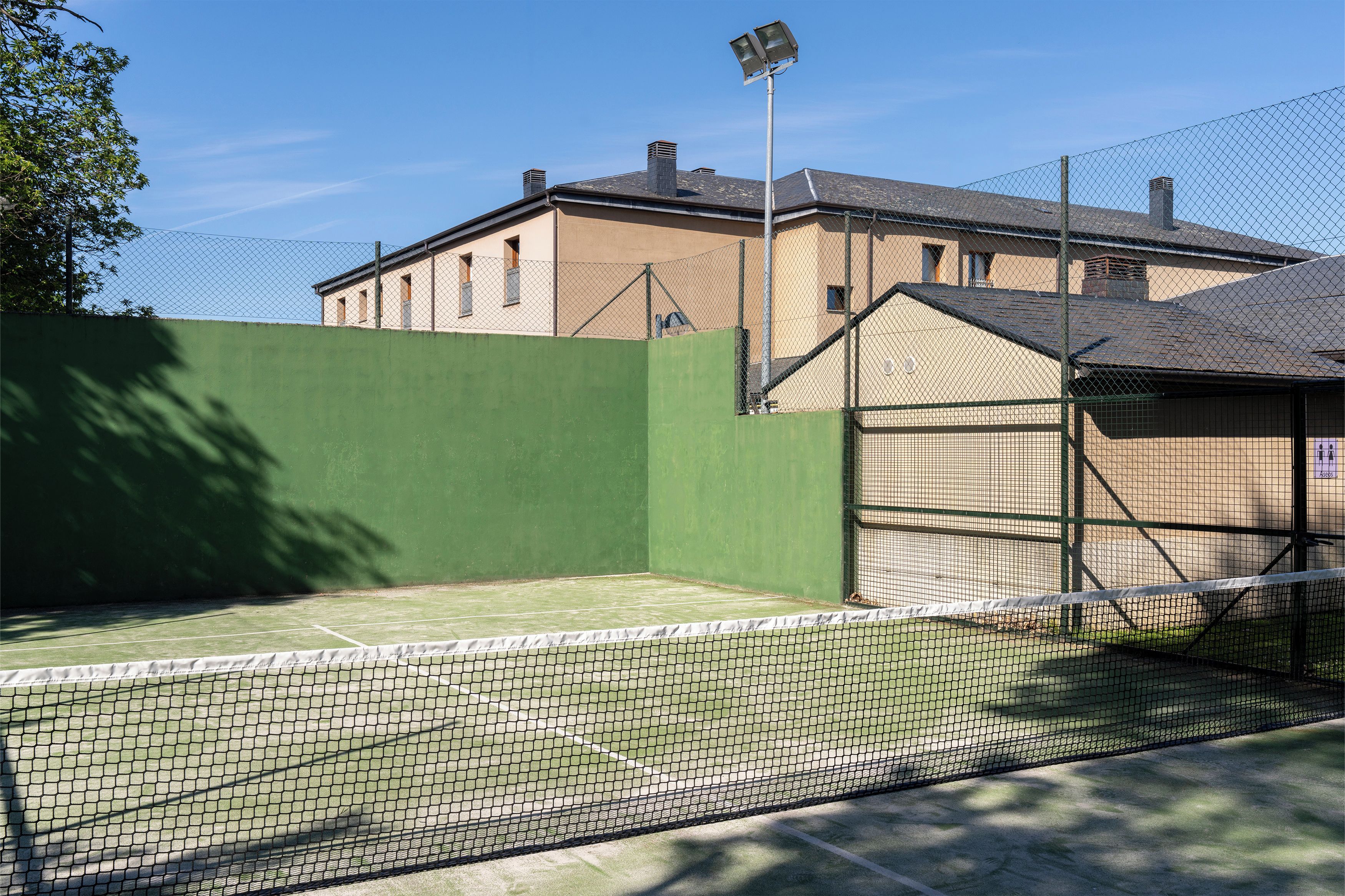 Una cancha de tenis con una red, rodeada por una cerca de malla metálica, y un edificio al fondo.