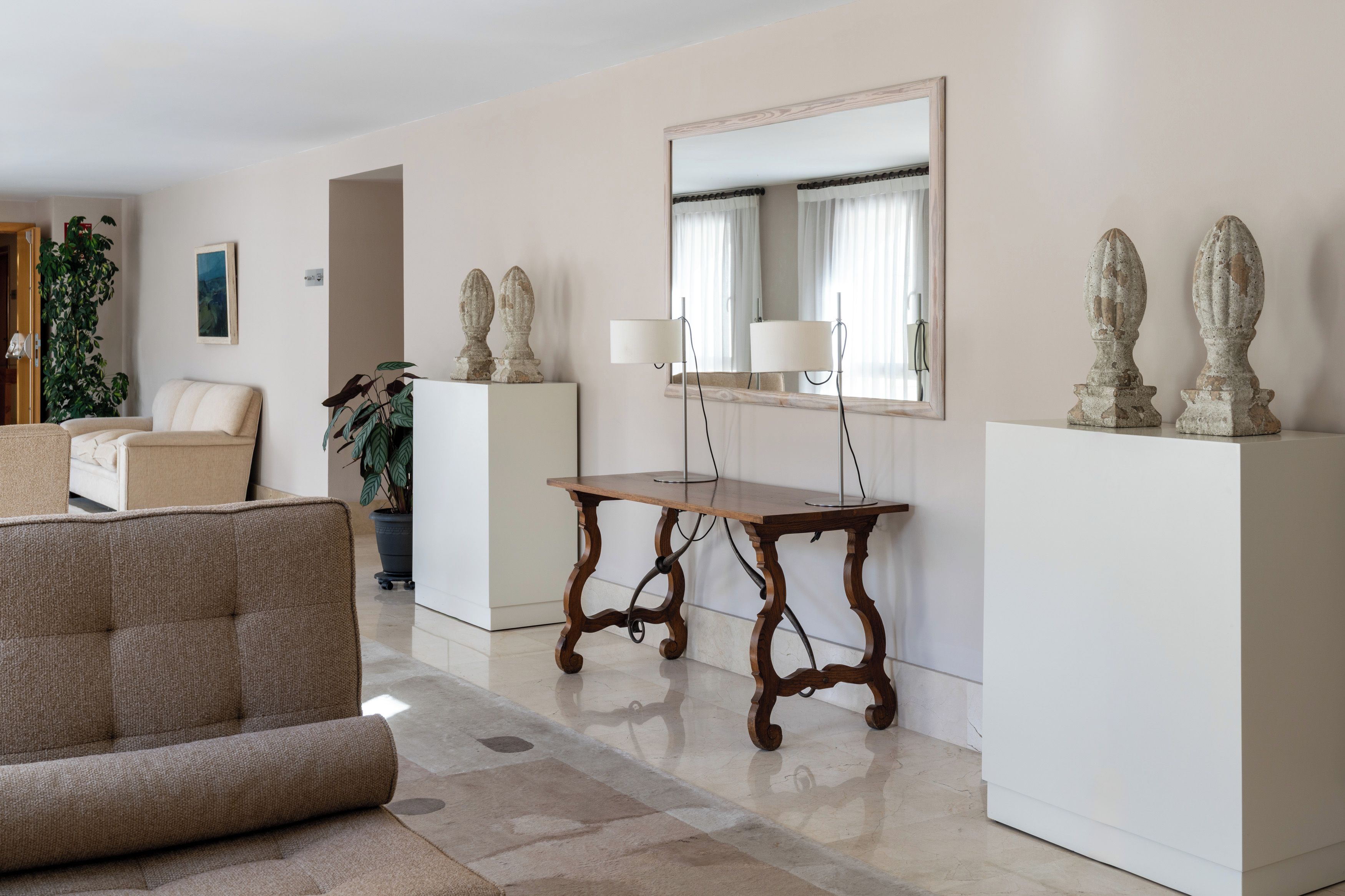 Una sala de estar con sofá, una mesa, un espejo y elementos decorativos sobre pedestales.