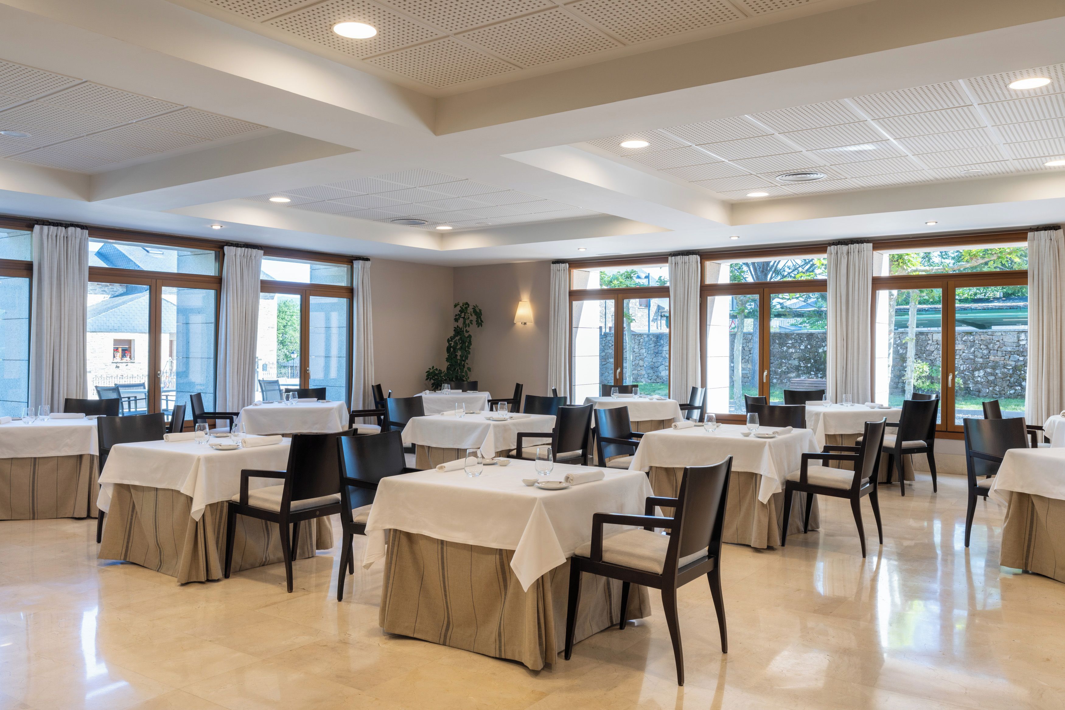 Un comedor amplio con varias mesas y sillas, ventanas grandes con cortinas e iluminación empotrada en el techo.