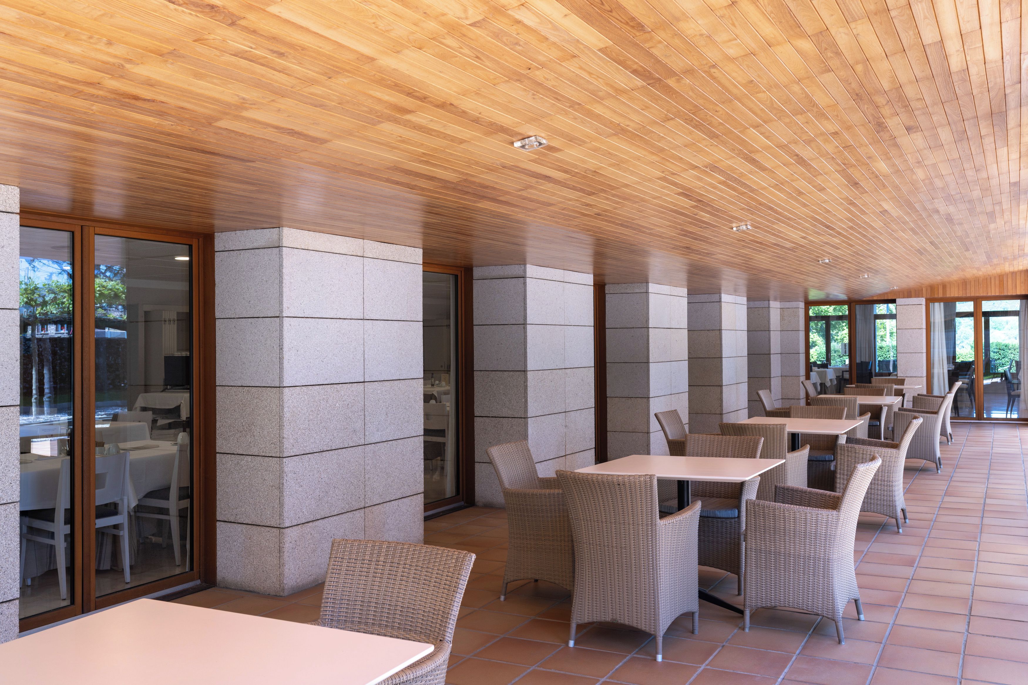 A covered outdoor seating area with tables and wicker chairs, stone pillars, and a wooden ceiling.