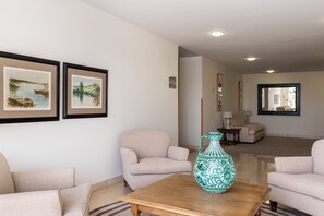 Una sala de estar con un sofá, sillones, una mesa de centro y un florero. Hay dos cuadros en la pared.