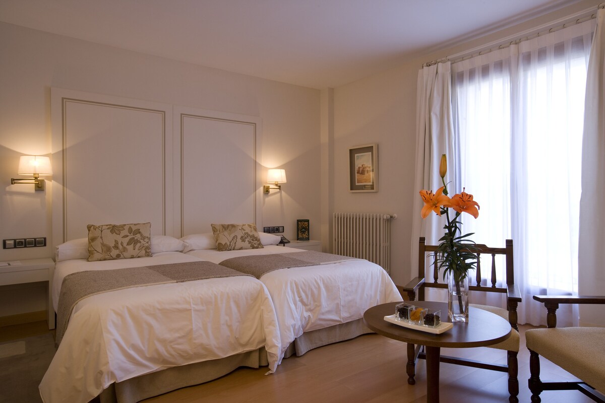 A hotel room with two beds, a small table with a vase of flowers, a chair, and a view of the outdoors through sheer curtains.