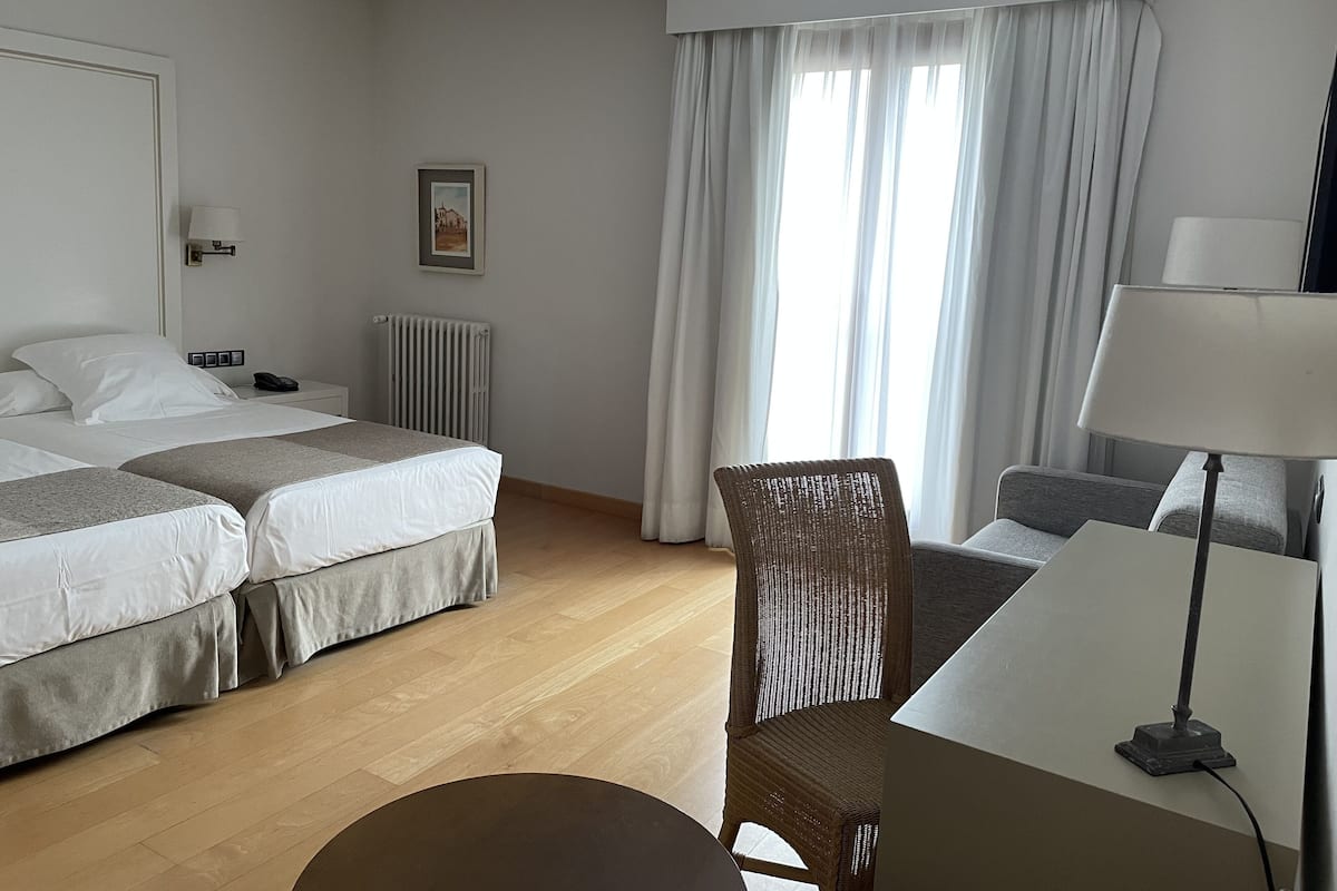 Une chambre d’hôtel avec deux lits, une chaise, une table, une lampe, un radiateur et une fenêtre avec des rideaux.
