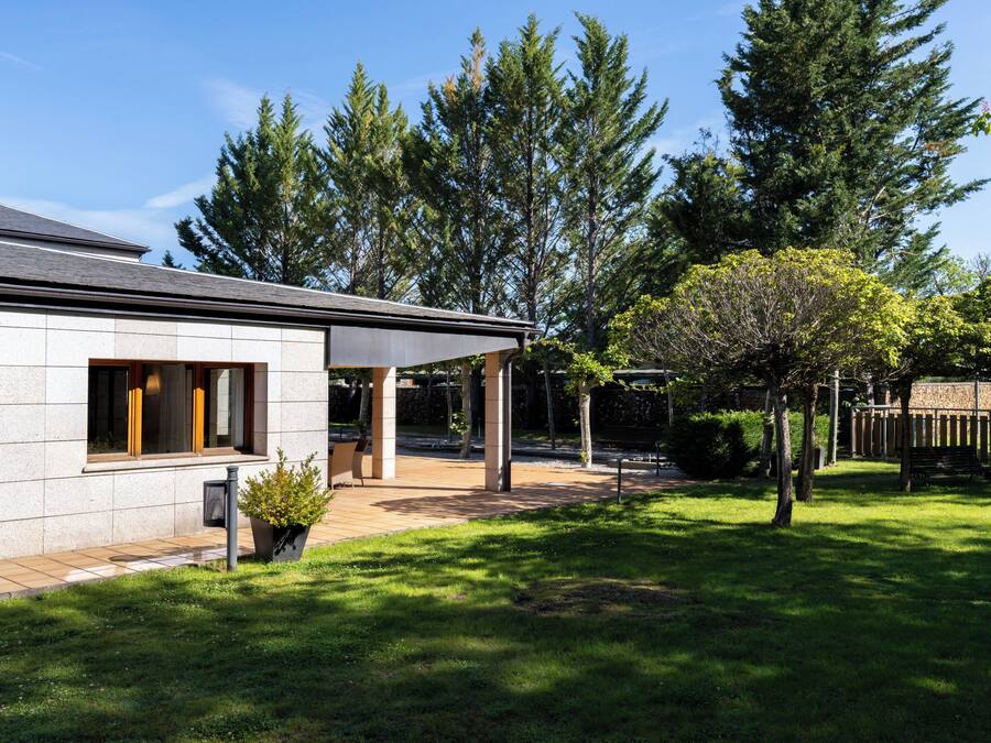 Una casa moderna con un porche cubierto, césped y altos árboles al fondo.