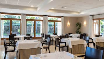 Un restaurante con varias mesas, sillas y amplias ventanas con cortinas.