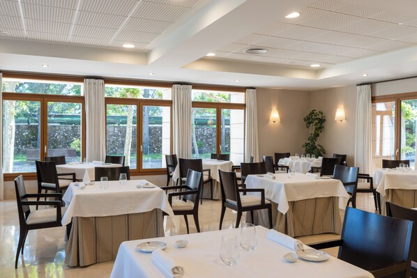Un restaurante con varias mesas, sillas y amplias ventanas con cortinas.