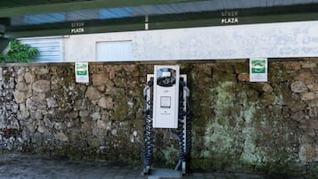 Un área de estacionamiento con estaciones de carga para vehículos eléctricos, un muro de piedra y un edificio con un cartel que dice 'PLAZA'.