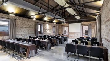 Un salón de conferencias con varias mesas y sillas, una pantalla de proyector y una pared de ladrillo.