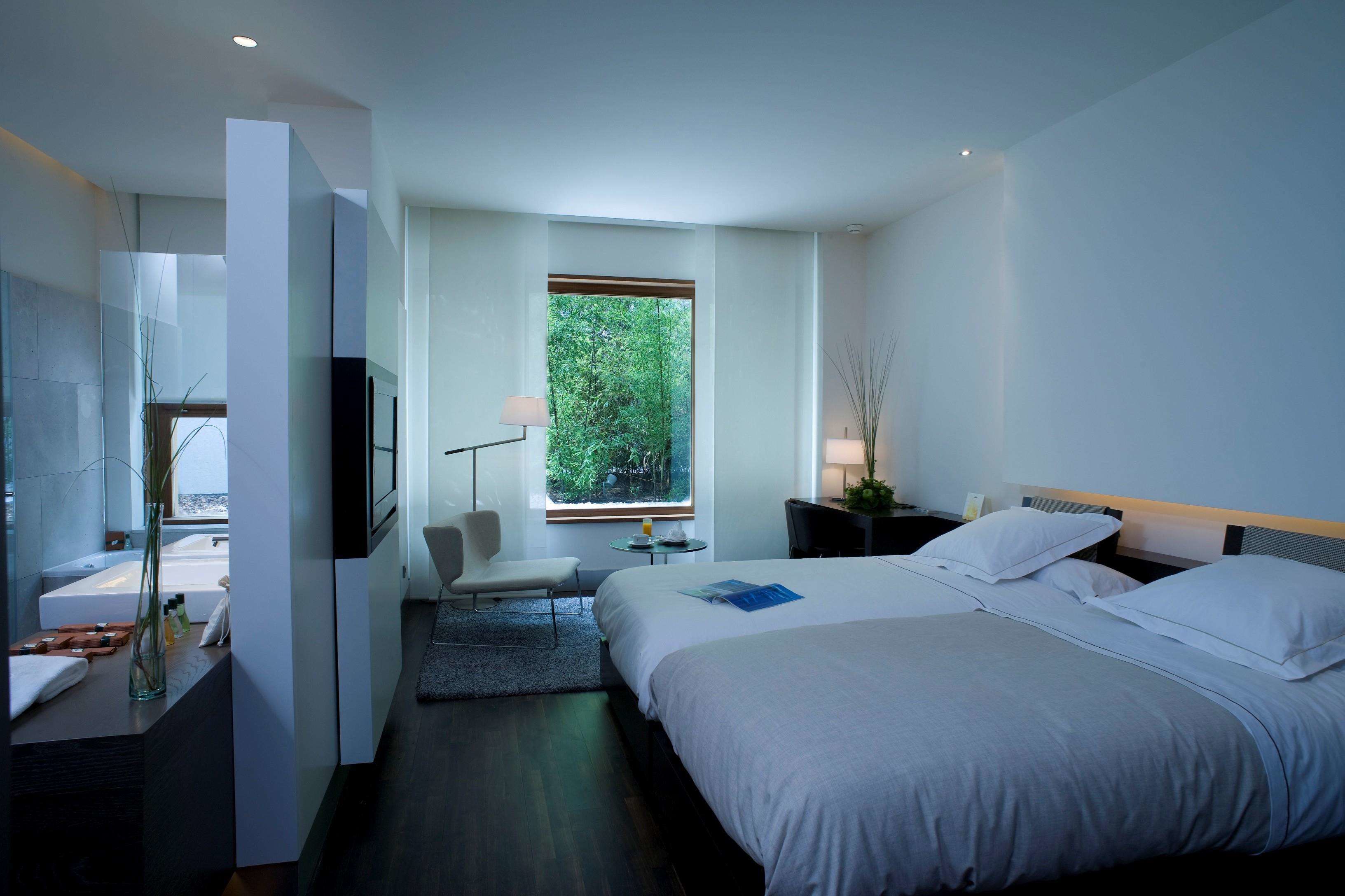 A modern hotel room with a large bed, a sitting area, a bathroom with a bathtub, and a large window offering a view of greenery.