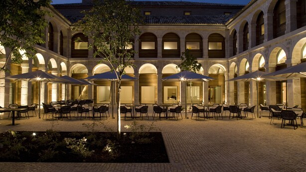 A courtyard with outdoor seating and umbrellas, surrounded by stone buildings with arched windows. - Parador De Alcala (Alcala de Henares)