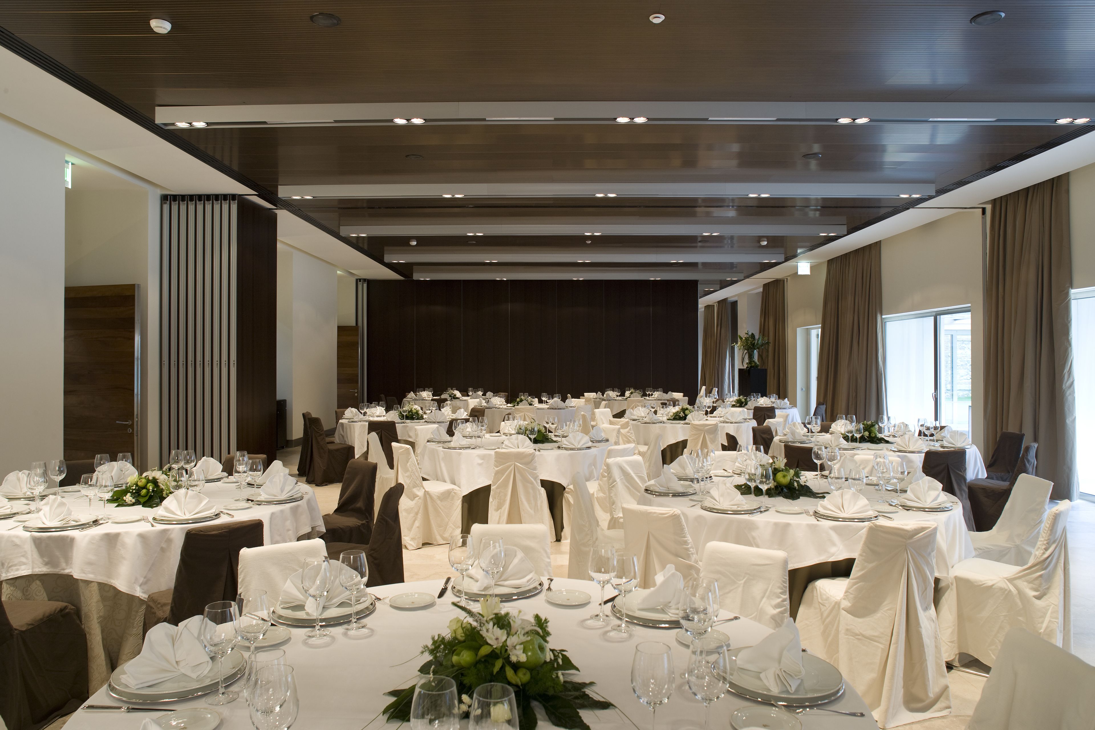Un gran saló de banquetes amb diverses taules rodones preparades per dinar, cadascuna amb estovatges blancs, cadira i elements decoratius al centre.
