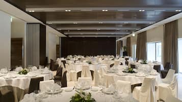 Un gran saló de banquetes amb diverses taules rodones preparades per dinar, cadascuna amb estovatges blancs, cadira i elements decoratius al centre.