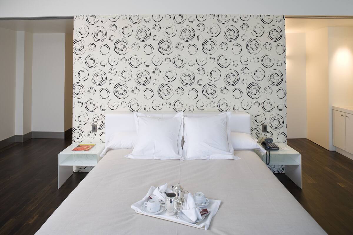 Une chambre d’hôtel moderne avec un grand lit, du linge de lit blanc, un plateau avec des articles pour le petit-déjeuner et un papier peint à motifs.
