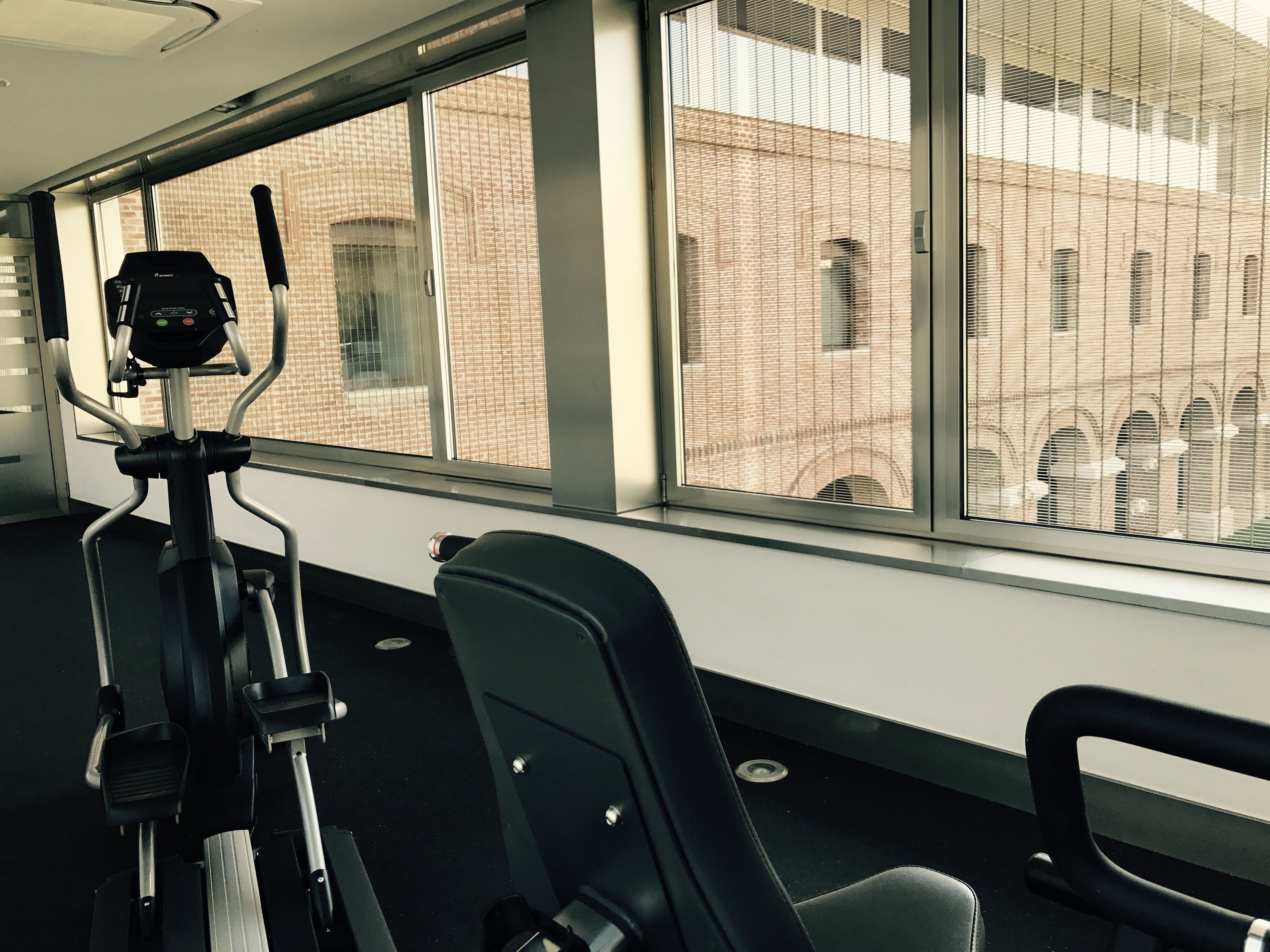 Un gimnasio con equipamiento de ejercicio, ventanas amplias y vista a un edificio de ladrillo.
