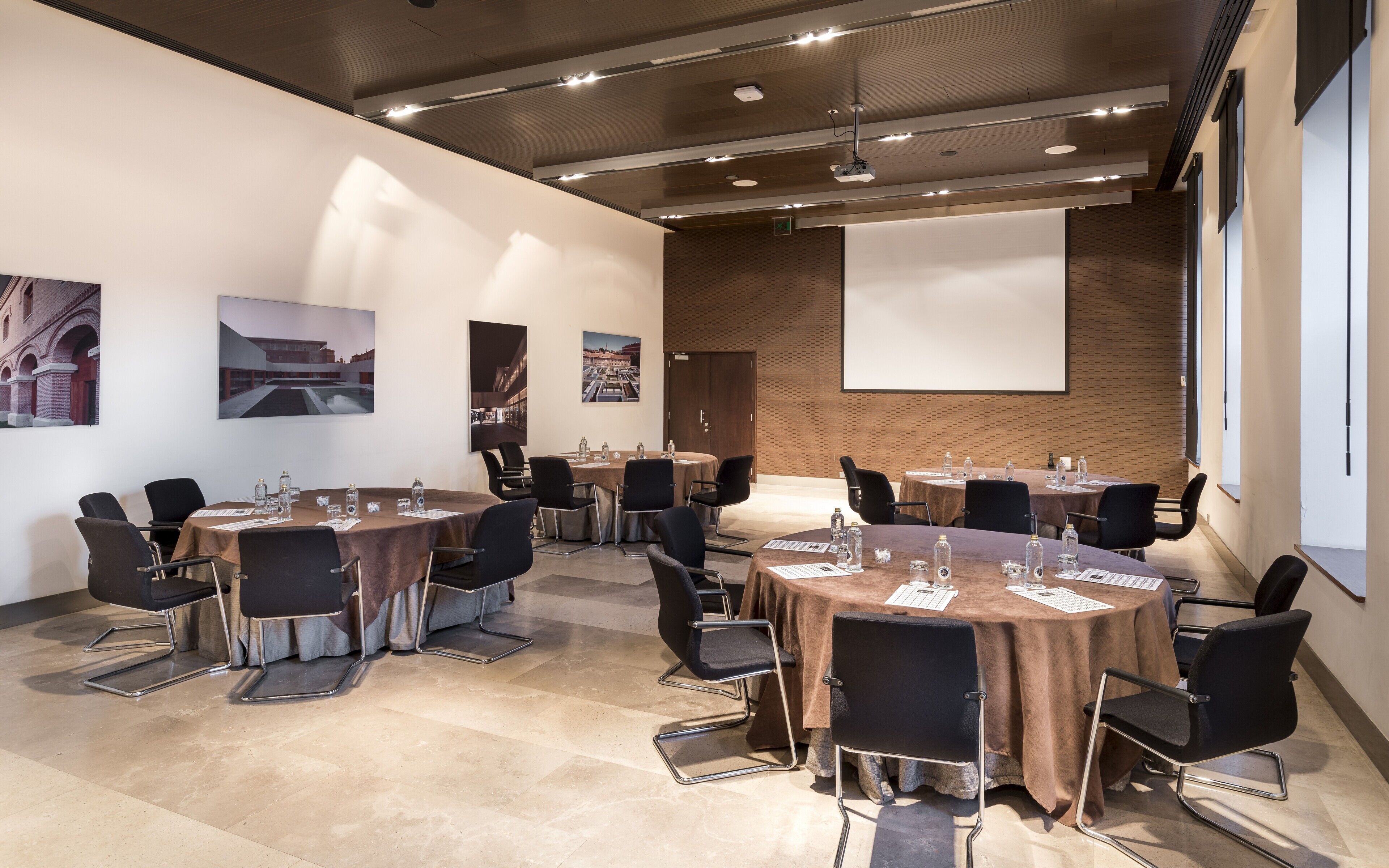 A conference room with multiple tables and chairs, equipped with water bottles and papers, in a modern building with large windows and a projector screen.