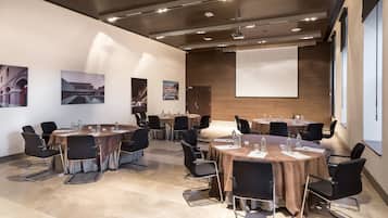 A conference room with multiple tables and chairs, equipped with water bottles and papers, in a modern building with large windows and a projector screen.