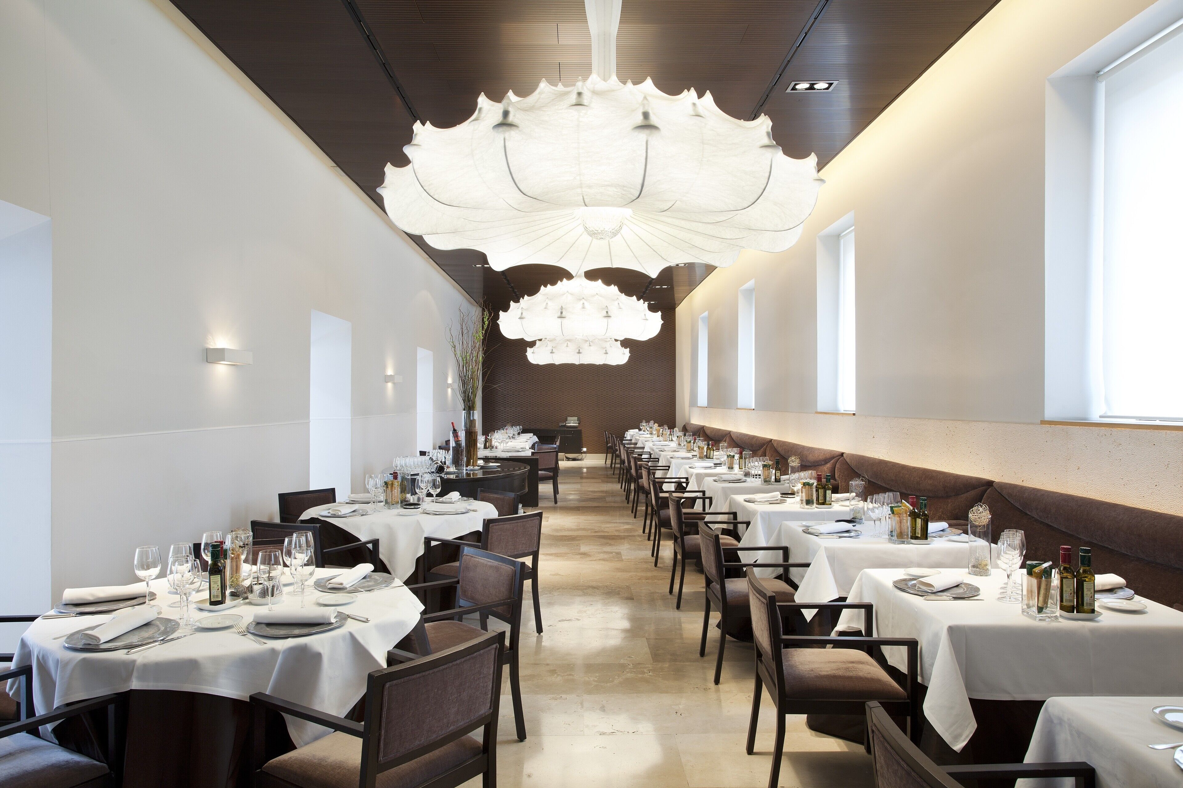 A long dining hall with multiple tables set for a meal, featuring a central chandelier and a row of windows along one wall.
