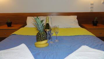 Una cama bien tendida con una colcha amarilla y azul, una piña, dos plátanos, una botella de champán y dos copas sobre la cama. Hay dos mesitas de noche con plantas en macetas a cada lado de la cama.