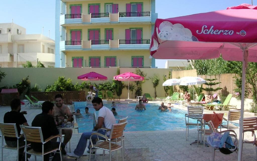 Un área de piscina con sillas de descanso y sombrillas, un edificio con balcones y un cartel que dice 'Scherzo'.