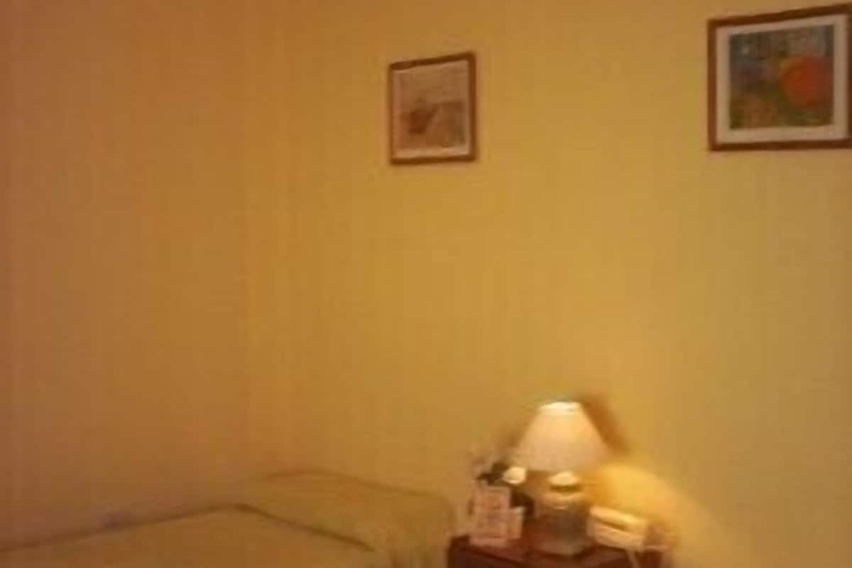 A small, dimly lit room with two single beds, a bedside table with a lamp, and two framed pictures on the wall.