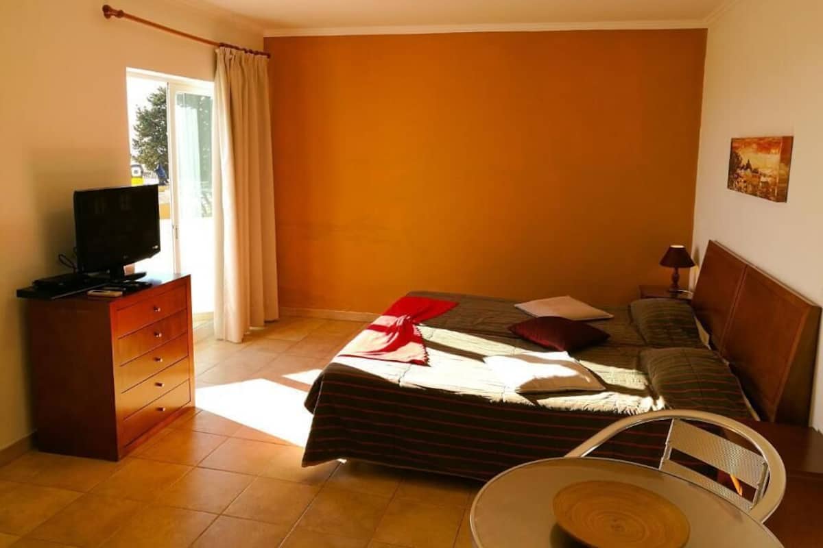 A bedroom with a bed, a television on a wooden dresser, a chair, and a painting on the wall.