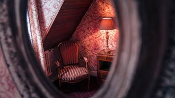 A room with a sloped ceiling, a chair, a table with a lamp, and floral wallpaper.