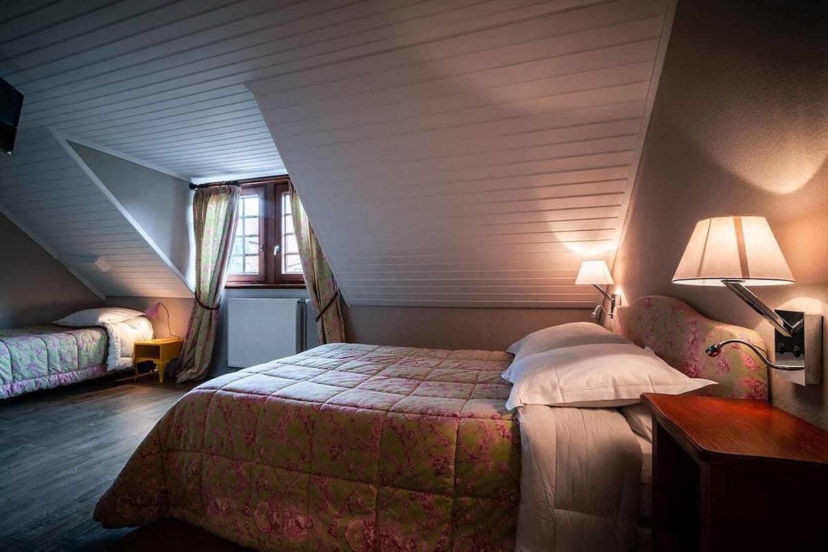 Quarto com teto inclinado, cama com colcha florida, mesa de cabeceira com abajur e janela com cortinas.