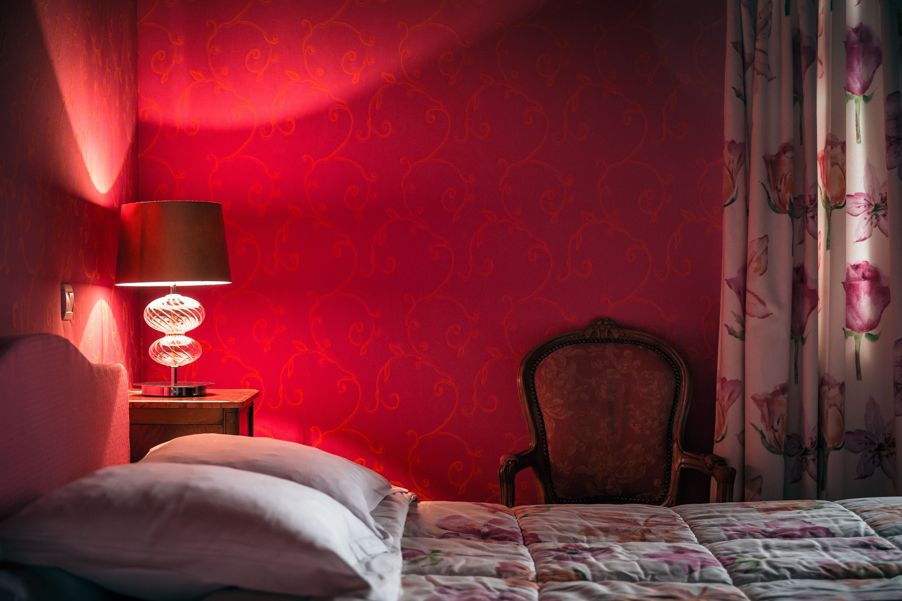 A bedroom with a red patterned wall, a bed with floral bedding, a wooden nightstand with a lamp, and a chair.