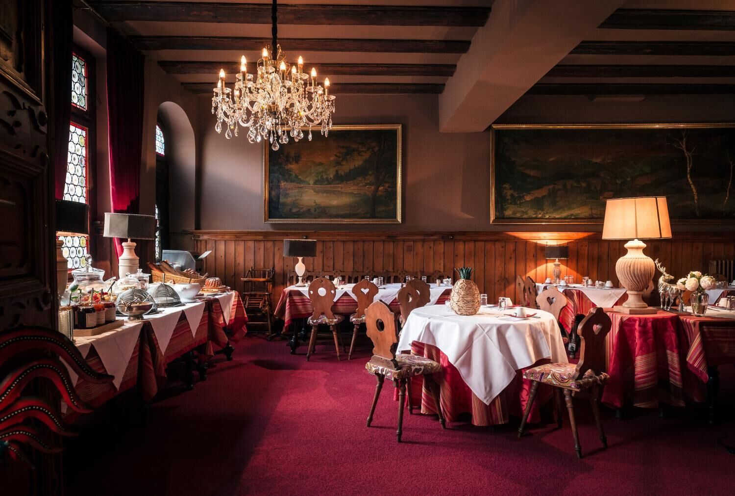 Een elegante eetzaal met houten lambrisering, een kroonluchter en rode karpetten waarop tafels zijn gedekt voor een maaltijd.