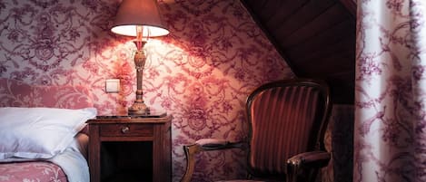 A bedroom with a bed, a nightstand with a lamp, a chair, and floral wallpaper.