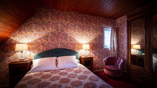 A bedroom with a sloped ceiling, floral wallpaper, a bed with white linens, two bedside lamps, a wooden nightstand, a leather armchair, and a large mirror. - Hotel Des Deux Clefs (Turckheim)