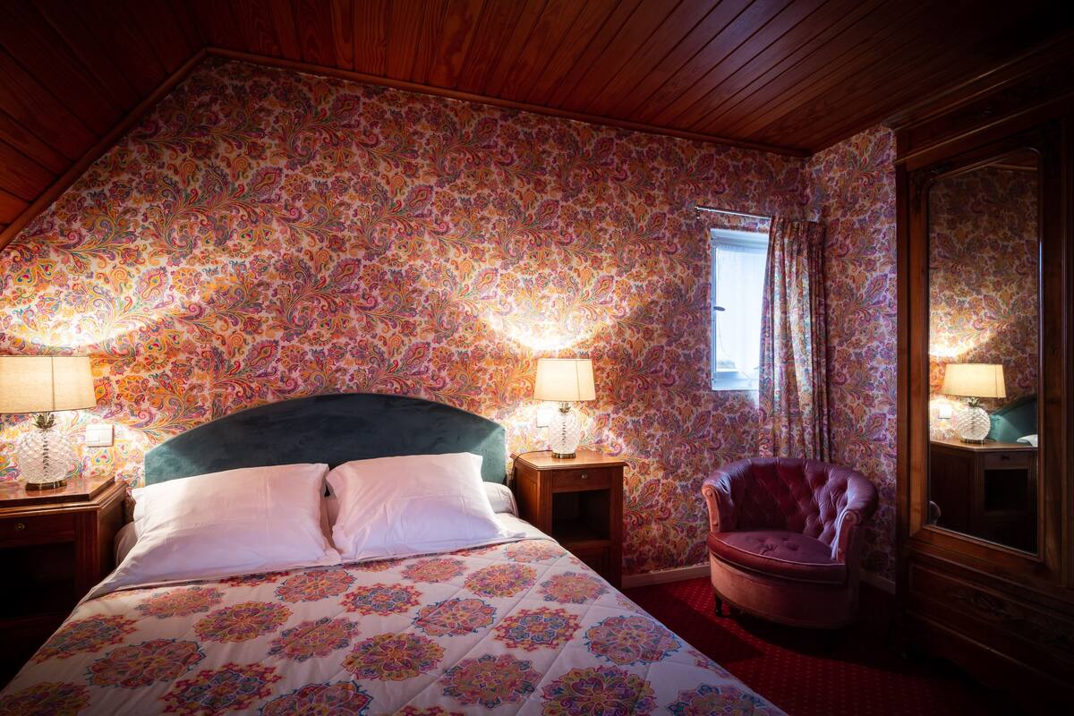 Quarto com teto inclinado, papel de parede floral, cama com roupa de cama branca, duas luminárias de cabeceira, mesa de cabeceira de madeira, poltrona de couro e um espelho grande.
