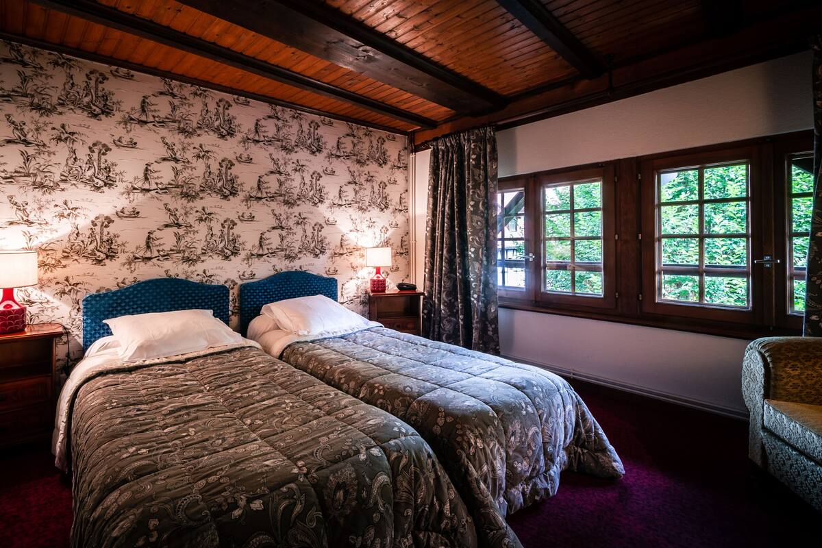 Quarto com duas camas, vigas de madeira no teto, papel de parede floral, janela com vista para vegetação e uma luminária vermelha em cada mesa de cabeceira.