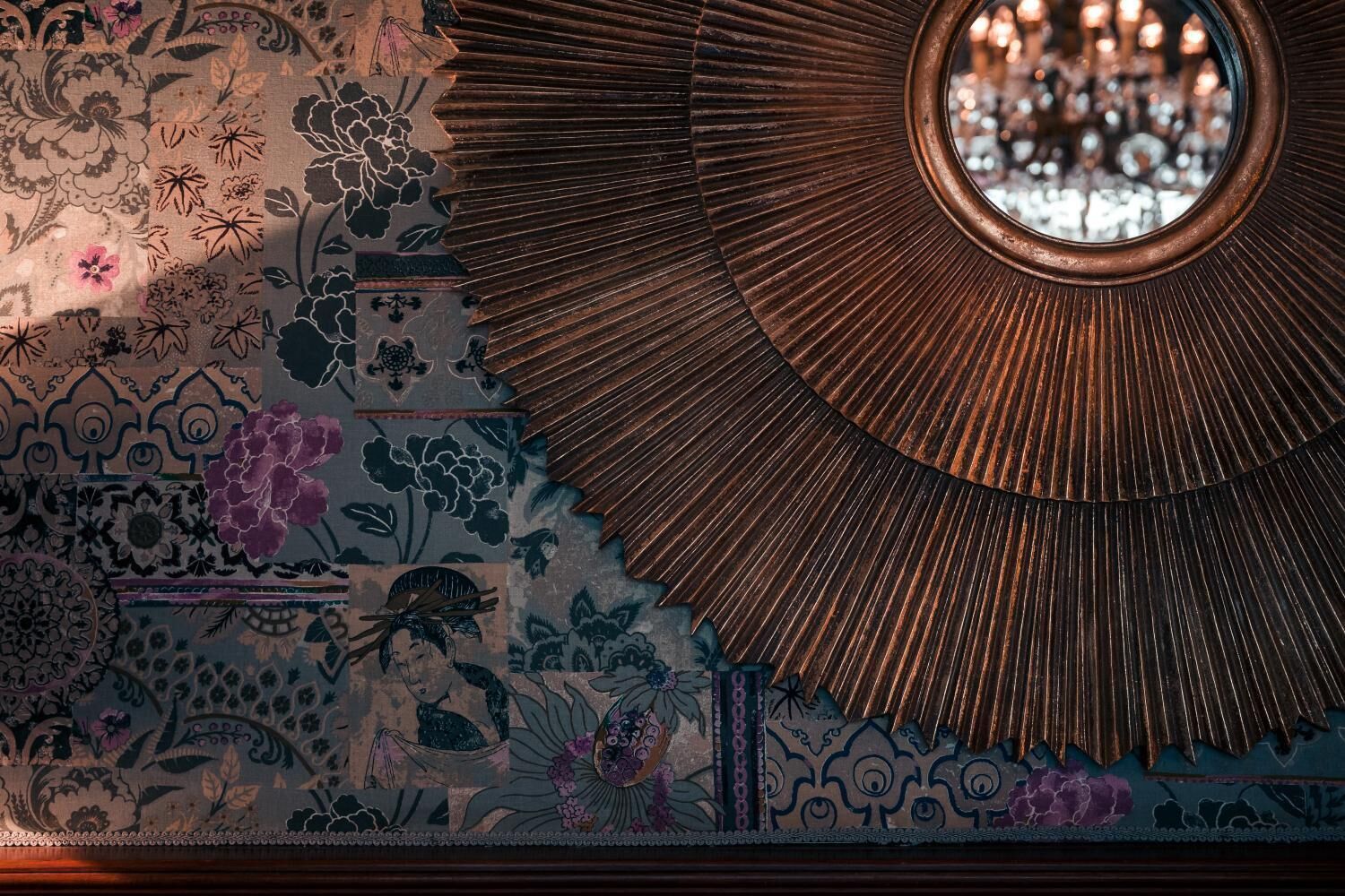 A detailed view of a room's wall with intricate floral and geometric patterns, a large circular mirror, and a chandelier.